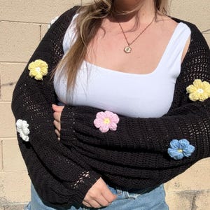 Puede incluir: Bolero de crochet negro adornado con aplicaciones florales tridimensionales de colores. Las flores son en tonos amarillo, rosa, blanco y azul. El bolero tiene mangas largas y un escote cuadrado. Se lleva sobre una camiseta sin mangas blanca.