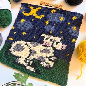May include: Crocheted square featuring a cow in a field under a starry night sky. The cow is white with black spots and has pink ears. The sky is blue with yellow stars and a yellow crescent moon. The field is green.