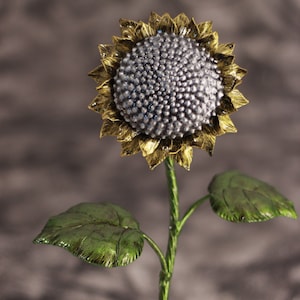Op de afbeelding: Een metalen zonnebloembeeldje van zilver en goud met groene bladeren. De zonnebloem heeft een getextureerd midden en de bladeren zien er realistisch uit.