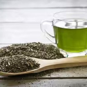 Può includere: Un cucchiaio di legno pieno di foglie di tè verde sfuse accanto a una tazza di vetro di tè verde su una superficie di legno bianca.