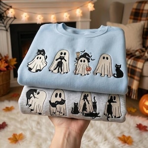 May include: Two crewneck sweatshirts stacked vertically. The top sweatshirt is light blue, featuring embroidered ghosts and black cats. The bottom sweatshirt is heather gray, also decorated with embroidered ghosts and black cats in various poses.