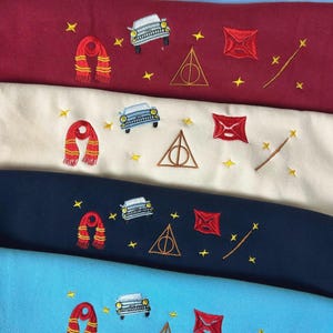 Puede incluir: Tejido bordado en tonos azul, burdeos, crema y marino. Cada pieza de tela presenta dise&ntilde;os bordados de una bufanda, un coche, un sobre, una varita y un s&iacute;mbolo triangular con un c&iacute;rculo en el interior, junto con estrellas amarillas.