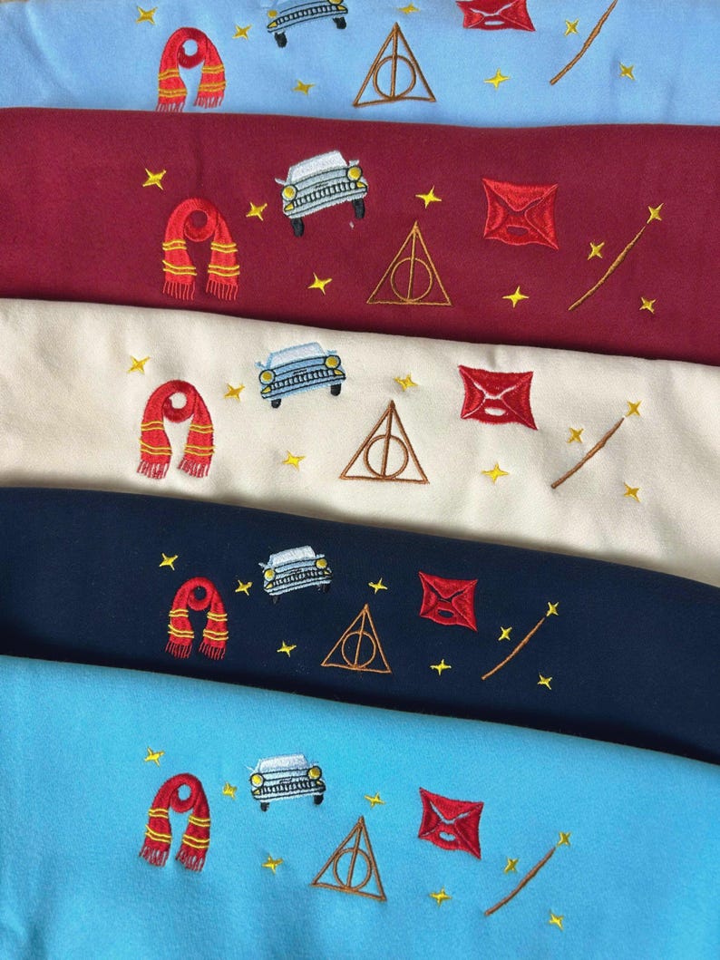 Puede incluir: Tejido bordado en tonos azul, burdeos, crema y marino. Cada pieza de tela presenta dise&ntilde;os bordados de una bufanda, un coche, un sobre, una varita y un s&iacute;mbolo triangular con un c&iacute;rculo en el interior, junto con estrellas amarillas.