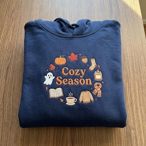 Pode incluir: Uma camisola com capuz azul-marinho com um design bordado com o texto "Cozy Season" ao centro. O bordado inclui uma abóbora, uma folha de ácer, uma bolota, uma meia, uma vela, um cachecol, uma camisola, uma chávena, um livro e um fantasma.