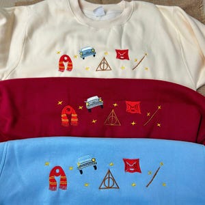 Puede incluir: Tres sudaderas en crema, burdeos y azul claro, cada una bordada con una bufanda, un coche, un tri&aacute;ngulo, un sobre, una varita y estrellas. Las sudaderas est&aacute;n apiladas, mostrando los dise&ntilde;os bordados.