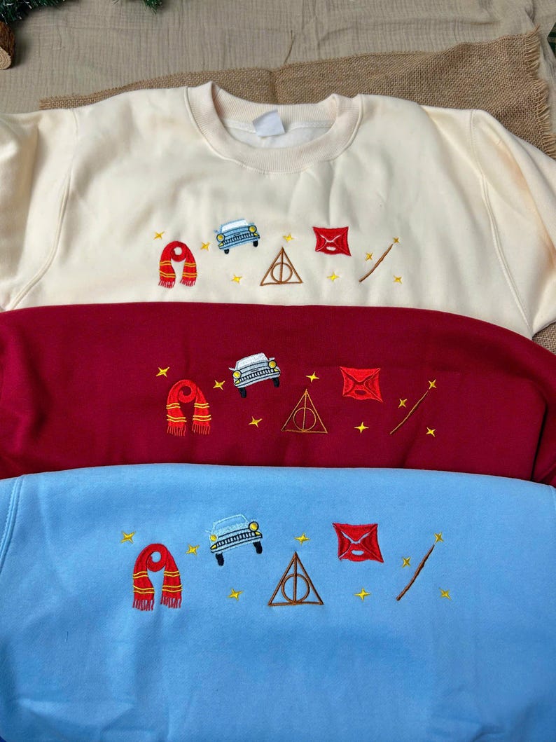 Puede incluir: Tres sudaderas en crema, burdeos y azul claro, cada una bordada con una bufanda, un coche, un tri&aacute;ngulo, un sobre, una varita y estrellas. Las sudaderas est&aacute;n apiladas, mostrando los dise&ntilde;os bordados.