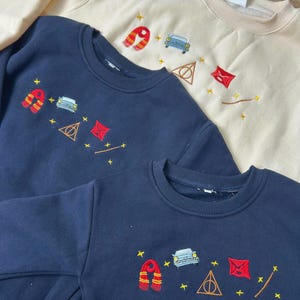 Puede incluir: Tres sudaderas de cuello redondo en azul marino y crema. Cada sudadera presenta dise&ntilde;os bordados, incluyendo una bufanda roja y dorada, un coche azul, un tri&aacute;ngulo dorado y un cuadrado rojo. Ropa para ni&ntilde;os.