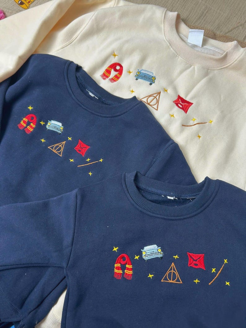 Puede incluir: Tres sudaderas de cuello redondo en azul marino y crema. Cada sudadera presenta dise&ntilde;os bordados, incluyendo una bufanda roja y dorada, un coche azul, un tri&aacute;ngulo dorado y un cuadrado rojo. Ropa para ni&ntilde;os.