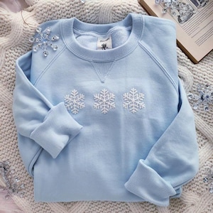 May include: A light blue sweatshirt with three embroidered white snowflakes. The long-sleeved top is folded on a white knitted blanket. A book and decorative snowflake ornaments are also visible.