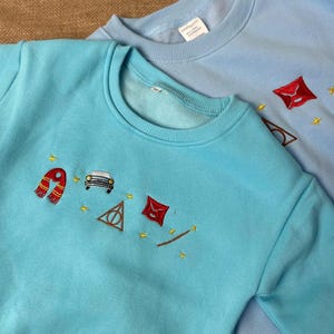 Puede incluir: Dos sudaderas azul claro con dise&ntilde;os bordados. Una es de un azul m&aacute;s brillante con una bufanda roja, un coche, un tri&aacute;ngulo y un sobre. La otra es de un azul m&aacute;s claro con un sobre, gafas y estrellas. Ambas tienen cuello redondo.