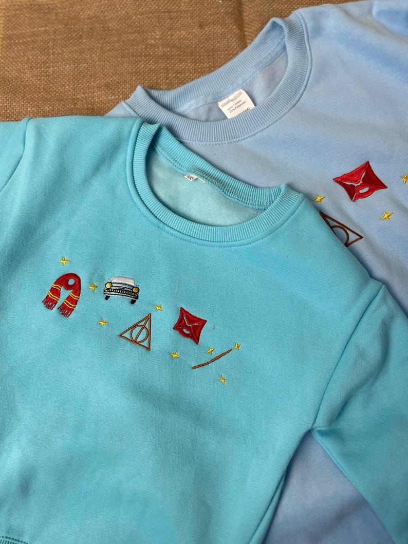 Puede incluir: Dos sudaderas azul claro con dise&ntilde;os bordados. Una es de un azul m&aacute;s brillante con una bufanda roja, un coche, un tri&aacute;ngulo y un sobre. La otra es de un azul m&aacute;s claro con un sobre, gafas y estrellas. Ambas tienen cuello redondo.