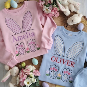 以下が含まれることがあります： ウサギの耳と足跡が刺繍されたパステルカラーのトレーナー2枚。ピンクのトレーナーにはアメリアの名前が、ブルーのトレーナーにはオリバーの名前が刺繍されています。