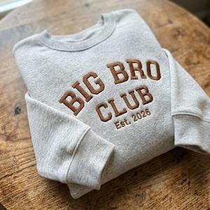 May include: A light gray crewneck sweatshirt featuring brown embroidered text that reads BIG BRO CLUB and Est. 2026. The garment has long sleeves with ribbed cuffs and a ribbed neckline, showcasing a textured fabric finish throughout the design.