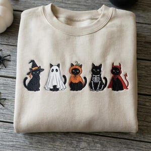Puede incluir: Una sudadera beige de cuello redondo con un diseño bordado de cinco gatos negros con varios disfraces de Halloween, incluyendo una bruja, un fantasma, una calabaza, un esqueleto y un diablo, dispuestos en fila horizontal en el pecho.