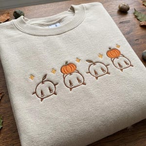 Peut inclure: Un sweat-shirt à col rond beige orné d'une broderie représentant quatre personnages ronds semblables à des fantômes. Deux de ces personnages portent des citrouilles orange sur la tête, avec de petites étoiles jaunes brodées entre eux.