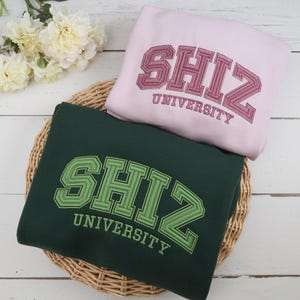May include: Two sweatshirts with the text "SHIZ UNIVERSITY" embroidered on the front. The sweatshirt on top is pink with burgundy embroidery. The sweatshirt on the bottom is dark green with green embroidery.