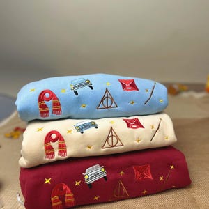 Puede incluir: Tres sudaderas bordadas en azul claro, crema y burdeos. Cada una presenta una bufanda, un coche, un rayo y un tri&aacute;ngulo con un c&iacute;rculo. Las sudaderas est&aacute;n apiladas una encima de otra.