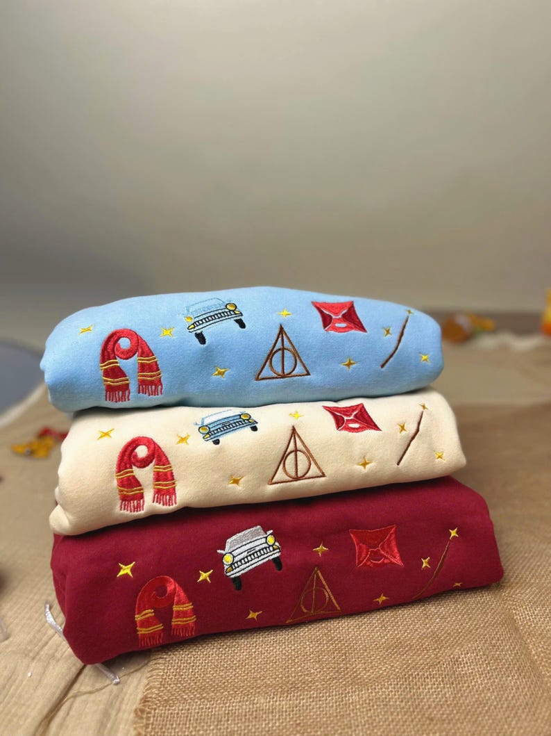 Puede incluir: Tres sudaderas bordadas en azul claro, crema y burdeos. Cada una presenta una bufanda, un coche, un rayo y un tri&aacute;ngulo con un c&iacute;rculo. Las sudaderas est&aacute;n apiladas una encima de otra.