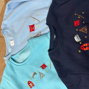 Puede incluir: Tres sudaderas de cuello redondo en azul claro, turquesa y azul marino. Cada sudadera presenta dise&ntilde;os bordados de una varita, una cometa roja, un tri&aacute;ngulo, un coche, una bufanda y estrellas. Las sudaderas est&aacute;n hechas de un material suave.