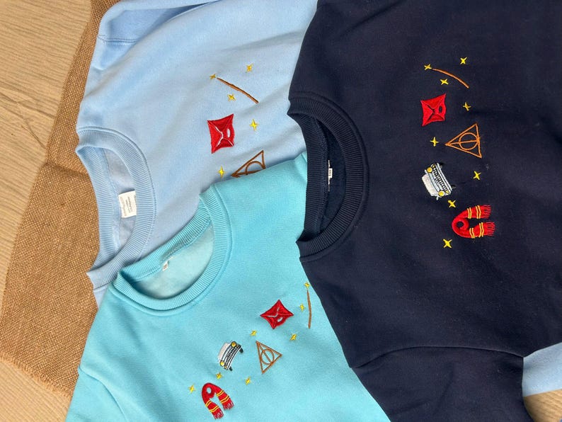 Puede incluir: Tres sudaderas de cuello redondo en azul claro, turquesa y azul marino. Cada sudadera presenta dise&ntilde;os bordados de una varita, una cometa roja, un tri&aacute;ngulo, un coche, una bufanda y estrellas. Las sudaderas est&aacute;n hechas de un material suave.