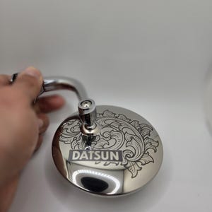 May include: A chrome-plated rearview mirror with a decorative floral design and the word "DATSUN" engraved on the surface.