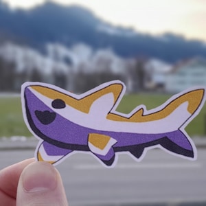 Puede incluir: Una pegatina de tiburón morado, amarillo y blanco con una cara sonriente. La pegatina está sobre un fondo blanco.