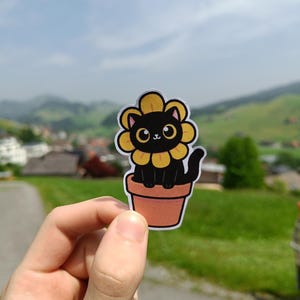 Könnte beinhalten: Ein Aufkleber mit einer schwarzen Katze mit Sonnenblumen-Design. Die Katze sitzt in einem braunen Blumentopf. Die Katze hat große Augen und einen niedlichen Ausdruck. Der Aufkleber wird vor einem Hintergrund aus grünem Gras und blauem Himmel gehalten.