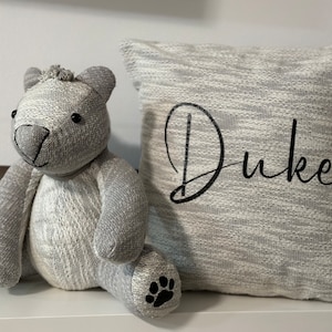 May include: A gray and white knitted plush teddy bear with black eyes and a black nose sits next to a gray and white knitted pillow with the name "Duke" embroidered in black.