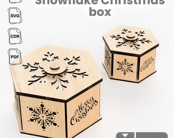 May include: Two wooden Snowflake Christmas boxes. The boxes are hexagonal with laser-cut snowflake designs on the lids and sides. The words "Merry Christmas" are written on the sides. The boxes are made of light-colored wood.