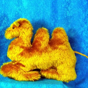 May include: A plush camel toy in shades of yellow and brown, with a textured, furry appearance. The camel has two humps, a long neck, and a small tail. The toy is set against a blue background.