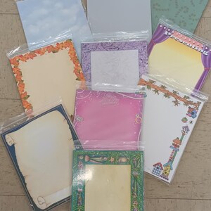 May include: A collection of decorative stationery sheets in clear plastic packaging. The sheets feature various designs, including floral borders, sky patterns, and stage curtains. The stationery is suitable for writing letters or notes.