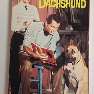 May include: A vintage VHS tape cover featuring a man and woman sitting with a large dog. The cover has the text "Disney Presents The Ugly Dachshund".