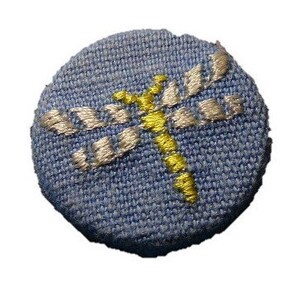 May include: A round, blue fabric patch with a dragonfly design. The dragonfly is embroidered in yellow and silver thread, with the wings and body clearly defined against the blue background. The patch is a small, decorative item.