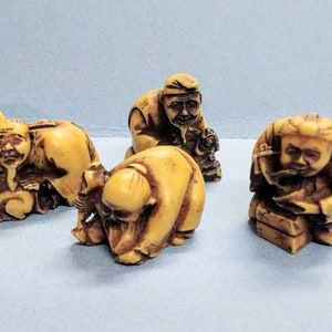 May include: A set of six small, intricately carved ivory figurines depicting various figures in traditional Japanese clothing. The figurines are in a light beige color with brown details and are arranged on a light blue surface.