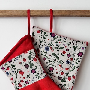Puede incluir: Un juego de guantes para horno rojos y blancos con un patrón floral. Los guantes están colgados de una barra de madera.