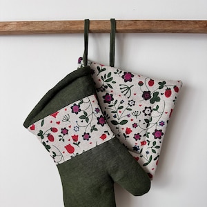 May include: A set of two green and white oven mitts with a floral pattern featuring red strawberries, pink and purple flowers, and green leaves. The mitts are hanging from a wooden bar.