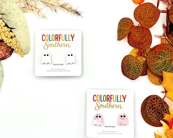 May include: Two pairs of ghost-shaped stud earrings on white display cards. One pair is solid white with black eyes, and the other pair features a marbled pink pattern with black eyes. Each card displays the text COLORFULLY SOUTHERN and Handmade.