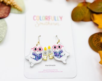 May include: A pair of white ghost-shaped earrings featuring pink glasses, holding blue glittery books and yellow candles. The earrings are displayed on a white card with the text Colorfully Southern, Handmade, and colorfullysouthernla.etsy.com.