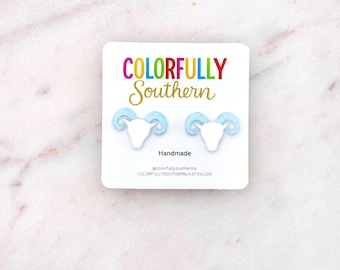 May include: A pair of stud earrings shaped like white ram heads with light blue glittery horns, mounted on a white display card featuring the text COLORFULLY Southern, Handmade, @colorfullysouthernla, and COLORFULLYSOUTHERNLA.ETSY.COM.
