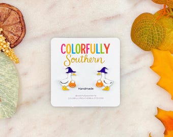 May include: A pair of white goose earrings featuring orange pumpkin buckets and purple witch hats on a white display card with the text COLORFULLY SOUTHERN and Handmade. The geese have yellow beaks and feet, and the card includes social media info.