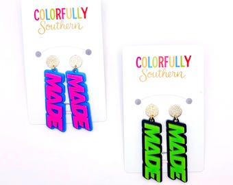 May include: Two pairs of dangle earrings on white display cards labeled Colorfully Southern. One pair features bright blue rectangular pendants with pink text reading MADE. The other pair has black rectangular pendants with neon green text reading MADE.