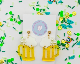 May include: A pair of gold earrings with a white foam beer mug design. The mugs are filled with a yellow liquid and have a white foam top with pearl accents. The earrings are surrounded by green, yellow, and blue sprinkles.