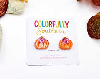May include: A pair of orange pumpkin-shaped stud earrings with glittery pink bows attached to the top. The earrings are mounted on a white display card featuring the text COLORFULLY SOUTHERN, Handmade, and the website address.