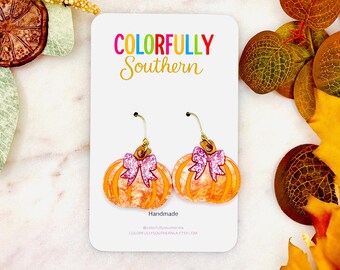 May include: A pair of orange pumpkin-shaped earrings on a white display card. Each pumpkin features a marbled orange acrylic finish, a gold-tone metal hook, and a small pink glitter bow attached to the top. The card displays the text COLORFULLY Southern.