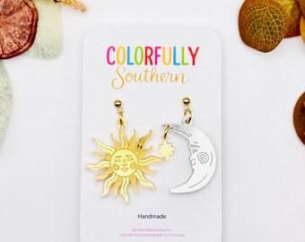 May include: A pair of mismatched earrings on a white display card labeled Colorfully Southern. One earring is a gold-toned sun with a face, and the other is a silver-toned crescent moon with a face and a small dangling star charm.