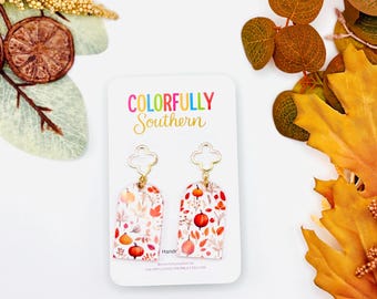 May include: A pair of arch-shaped dangle earrings featuring a white background with a colorful autumn pattern of orange pumpkins and fall leaves. The earrings hang from gold-toned clover-shaped posts on a white display card labeled Colorfully Southern.