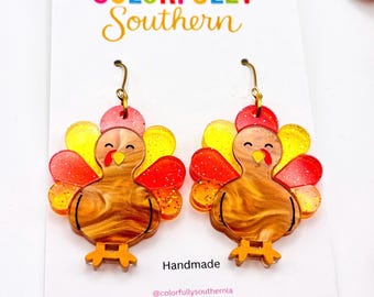 May include: A pair of dangle earrings shaped like turkeys with brown marbled bodies, yellow and red glittery tail feathers, and gold-toned hooks. The earrings are displayed on a white card featuring the text Colorfully Southern and Handmade.