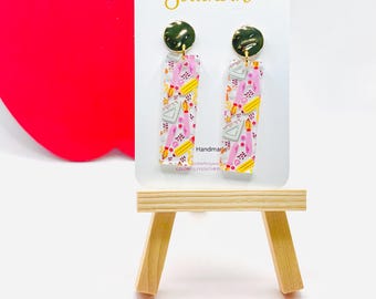 May include: A pair of rectangular dangle earrings featuring a school supply pattern with yellow pencils, pink paintbrushes, and gray rulers. The earrings have gold-tone circular studs and are displayed on a white card labeled Colorfully Southern.