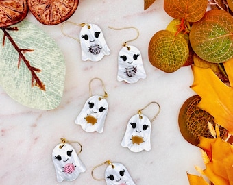 May include: Three pairs of white ghost-shaped earrings with gold-toned hooks. Each ghost features black eyes and a smile, holding a small object: a brown jar, a gold-colored pumpkin, or a pink yarn ball. The earrings have a textured surface.