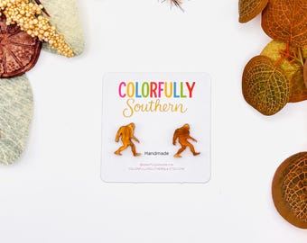 May include: A pair of amber-colored acrylic stud earrings shaped like Bigfoot figures, mounted on a white display card featuring the text COLORFULLY Southern, Handmade, and the website address COLORFULLYSOUTHERNLA.ETSY.COM in black text.